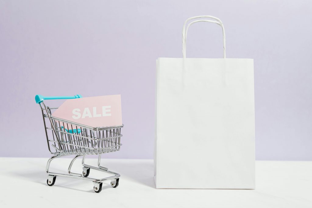 'sale' tag in a miniature shopping cart, next to a white shopping bag